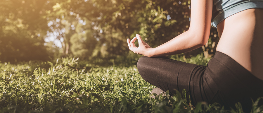 Person in meditation pose sitting on grass outdoors with sunlight filtering through trees