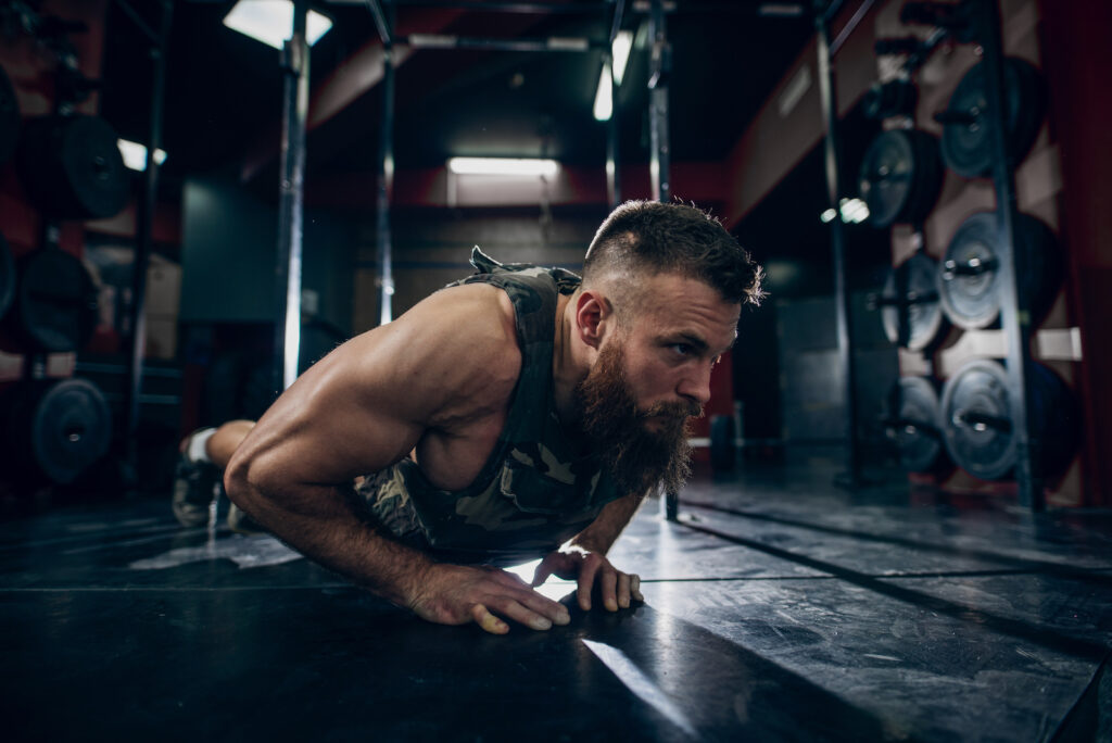 Bearded man performing a plank exercise on gym floor with weights visible in background