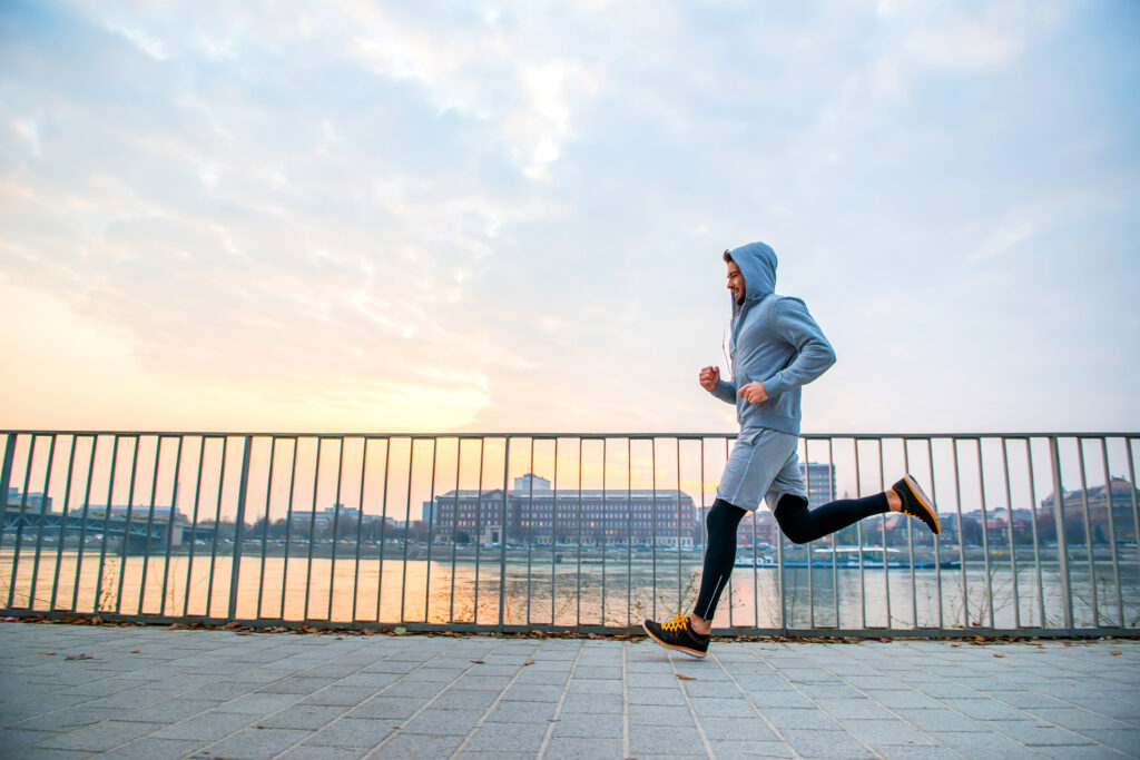 Person jogging along a waterfront promenade with city buildings and water visible behind a railing at sunset