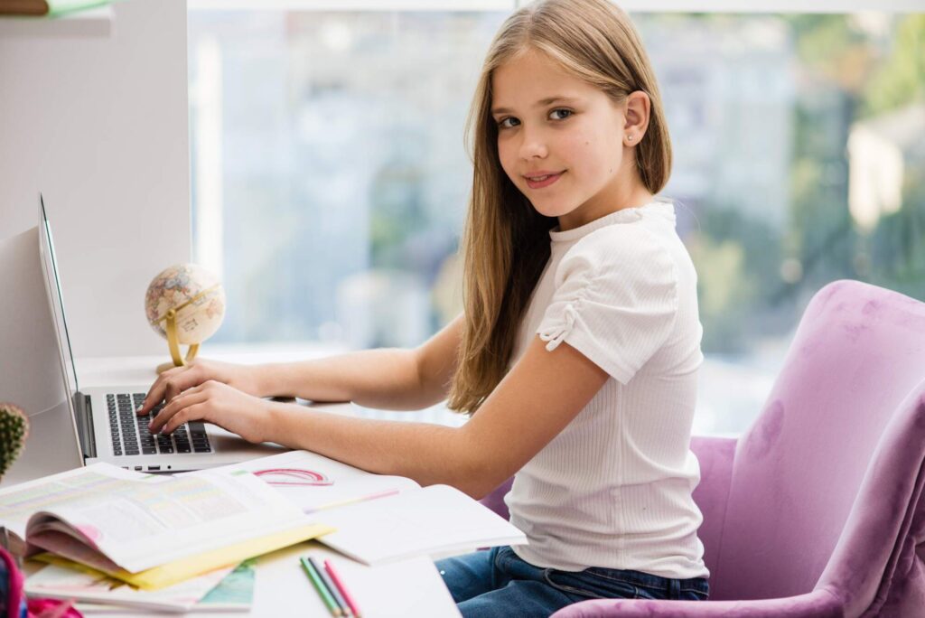 Young girl with long blonde hair sitting at a desk with a laptop, globe, and school supplies, looking over her shoulder at the camera with a slight smile