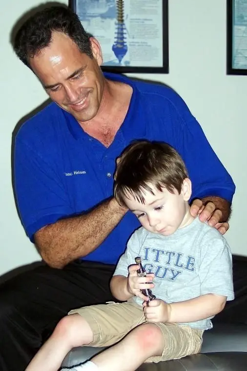Dr. Holmes, smiling, with his hands on the shoulders of a little boy in a “Little Guy” T-shirt sitting on the adjusting table