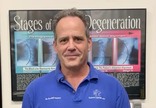Dr. Randall Holmes in a blue Holmes Chiropractic polo shirt, in front of a “Stages of Degeneration” spinal X-ray poster in his office