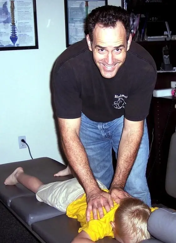 Dr. Holmes leaning over a boy lying face down on the adjusting table, his hands on the boy’s back, grinning at the camera